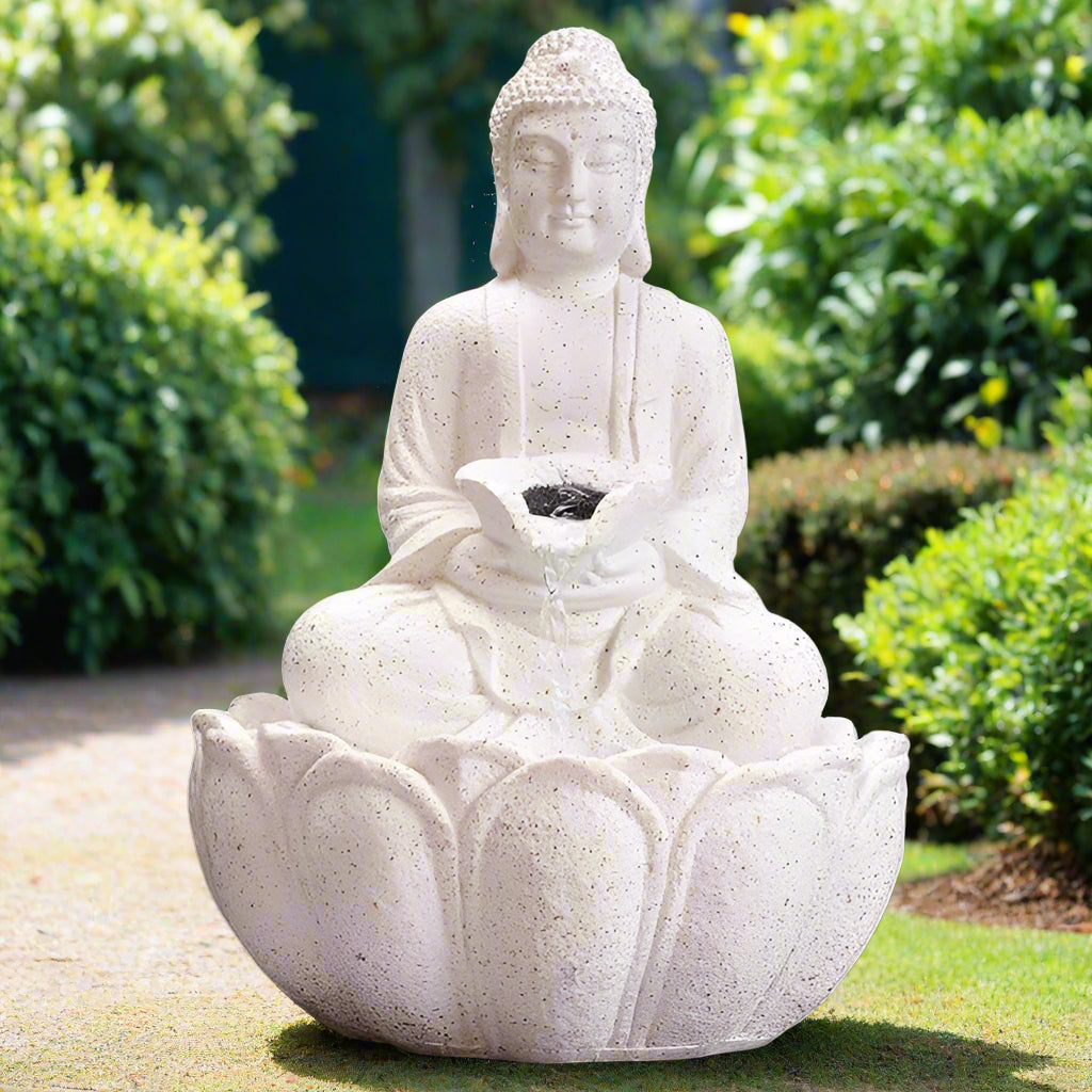 A white stone-looking Buddha water fountain with LED lighting placed on a wooden surface with greenery in the background.