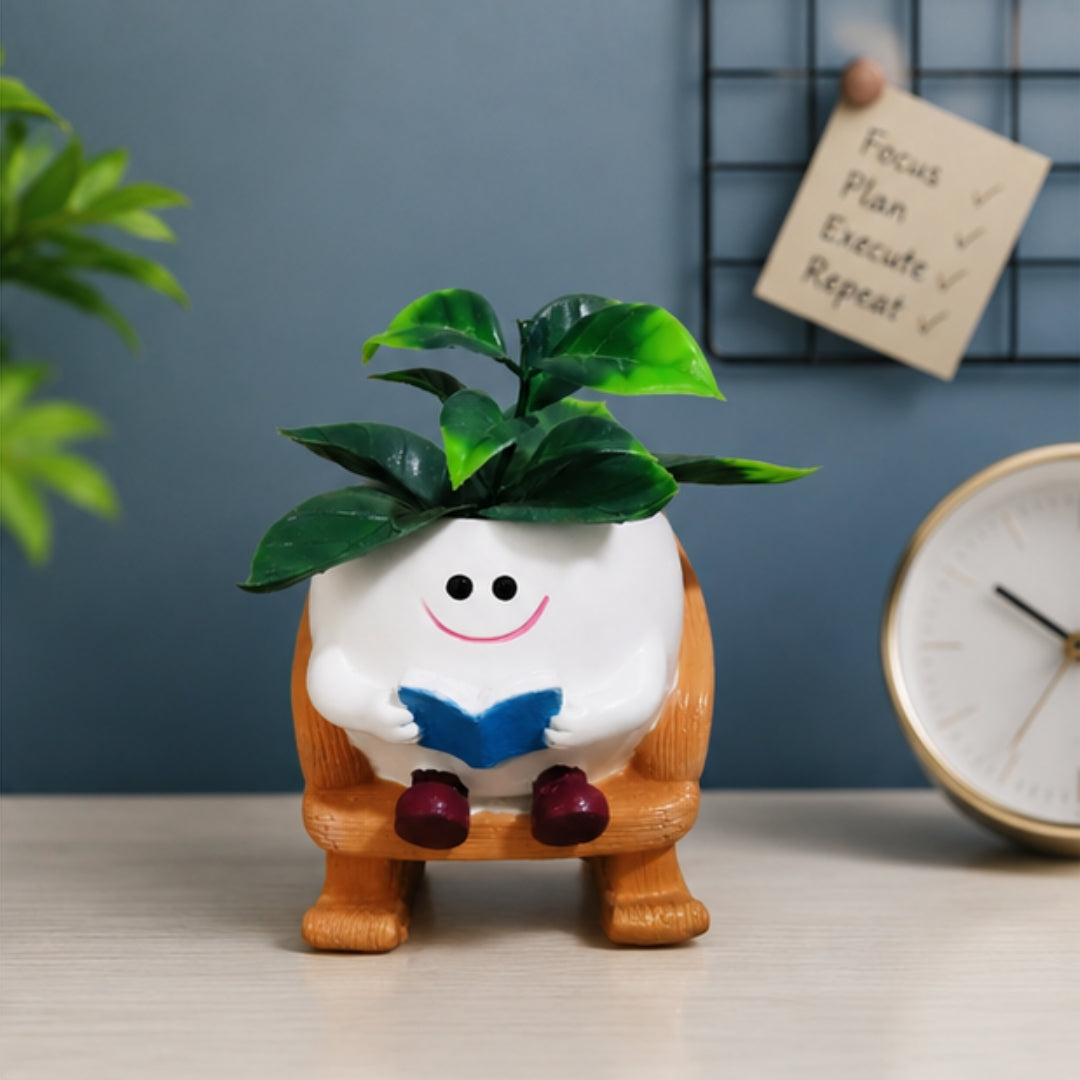 A small potted plant in a white round pot shaped like a smiling character sitting on an orange chair and reading a blue book, placed on a tabletop with a blue wall, plant, and clock in the background.