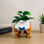 A small potted plant in a white round pot shaped like a smiling character sitting on a wooden chair, with green leaves on top, placed on a tabletop beside stacked books and another potted plant.