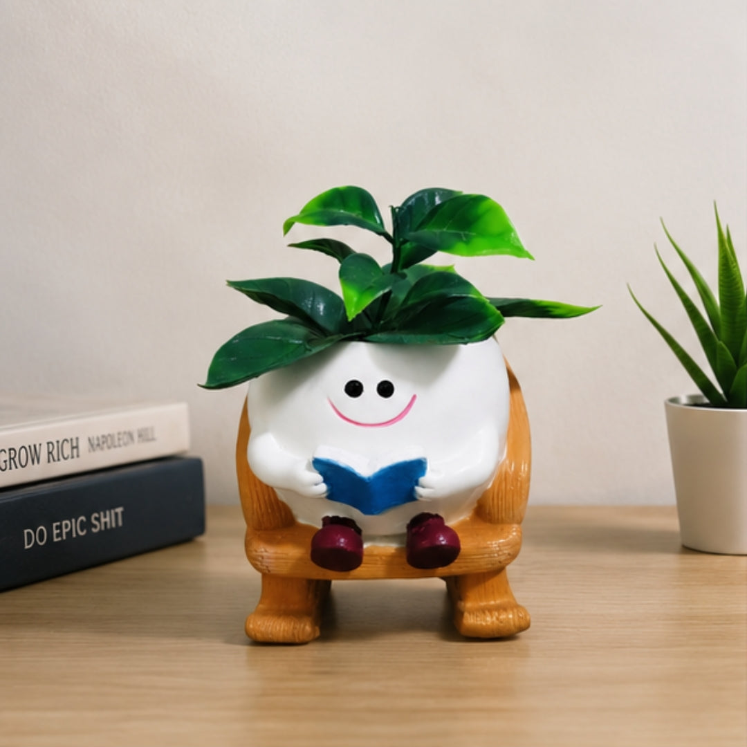 A small potted plant in a white round pot shaped like a smiling character sitting on a wooden chair, with green leaves on top, placed on a tabletop beside stacked books and another potted plant.