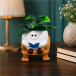 A ceramic plant pot shaped like a smiling white character sitting on a wooden chair and reading a blue book, with green leaves on top, displayed on a dark wooden table in a styled room setting.