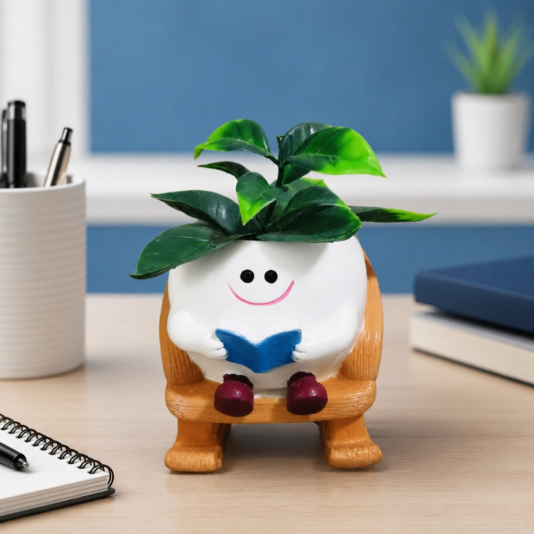A small decorative plant pot figurine shaped like a white smiling character sitting in a wooden chair and reading a blue book, with green leaves on top, displayed on a desk in a room setting.