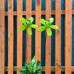 Two green flower-shaped garden wind spinners with cartoon eyes on thin stakes, shown in front of a wooden fence with a potted plant at the bottom.