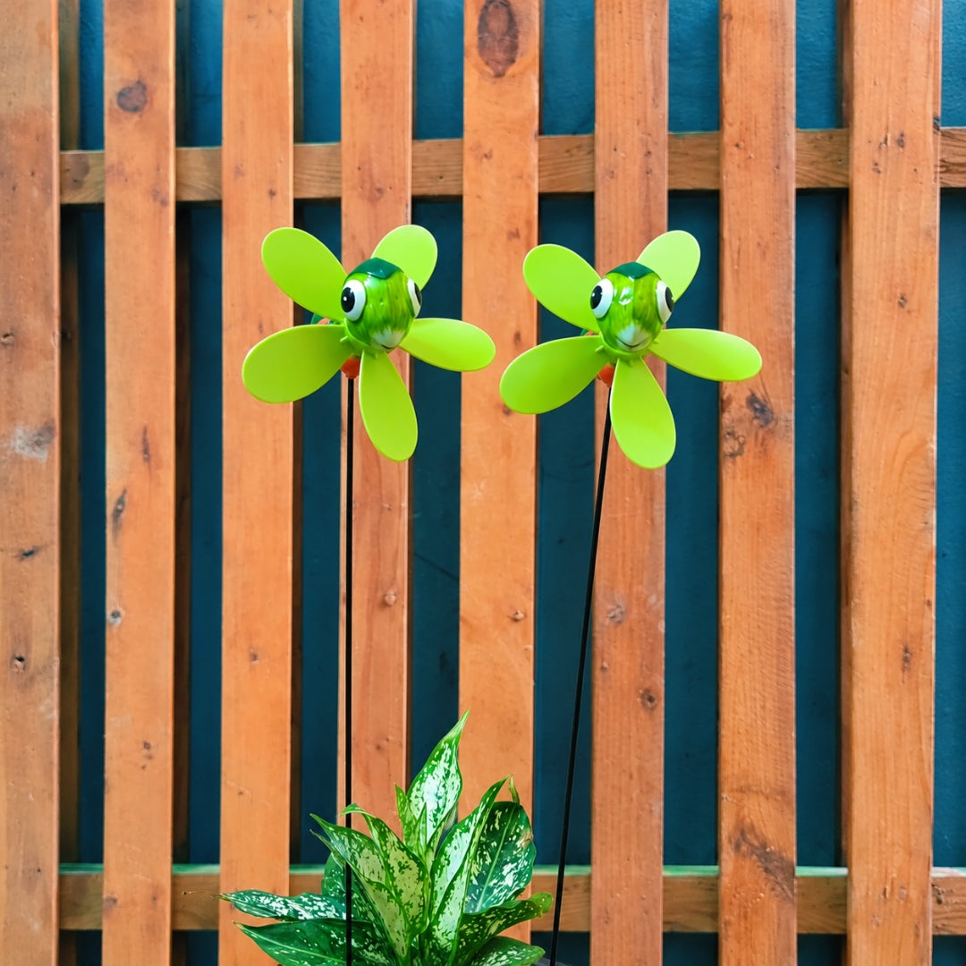 Two green flower-shaped garden wind spinners with cartoon eyes on thin stakes, shown in front of a wooden fence with a potted plant at the bottom.