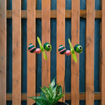 Two decorative wind-up insect garden lights shaped like green ladybugs with pink and black striped bodies and yellow-green propeller blades, mounted on black stakes in front of a wooden fence and above a potted plant.