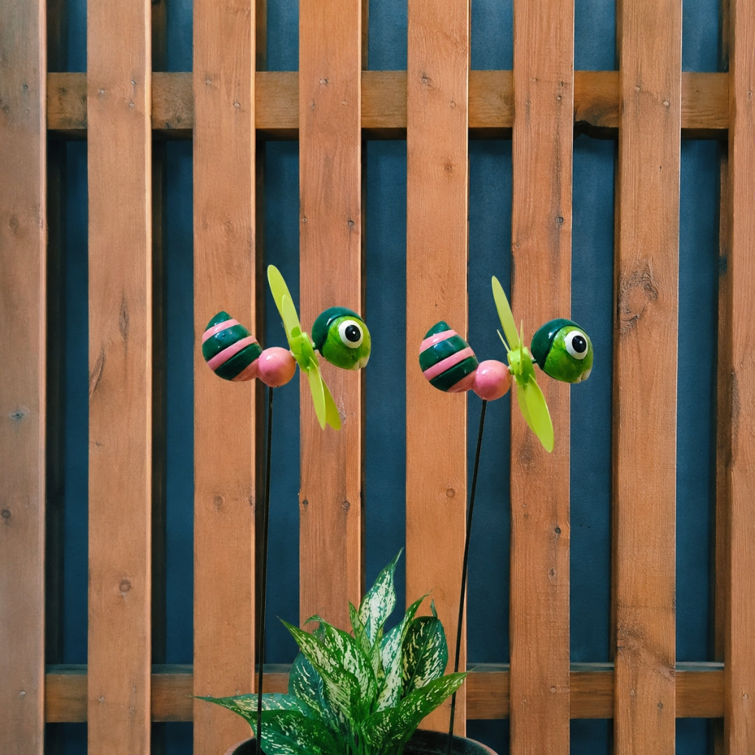 Two decorative wind-up insect garden lights shaped like green ladybugs with pink and black striped bodies and yellow-green propeller blades, mounted on black stakes in front of a wooden fence and above a potted plant.