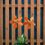 Two orange fish-shaped garden wind spinners with cartoon eyes mounted on black stakes, displayed in front of a brown wooden slat fence with a green plant at the bottom.