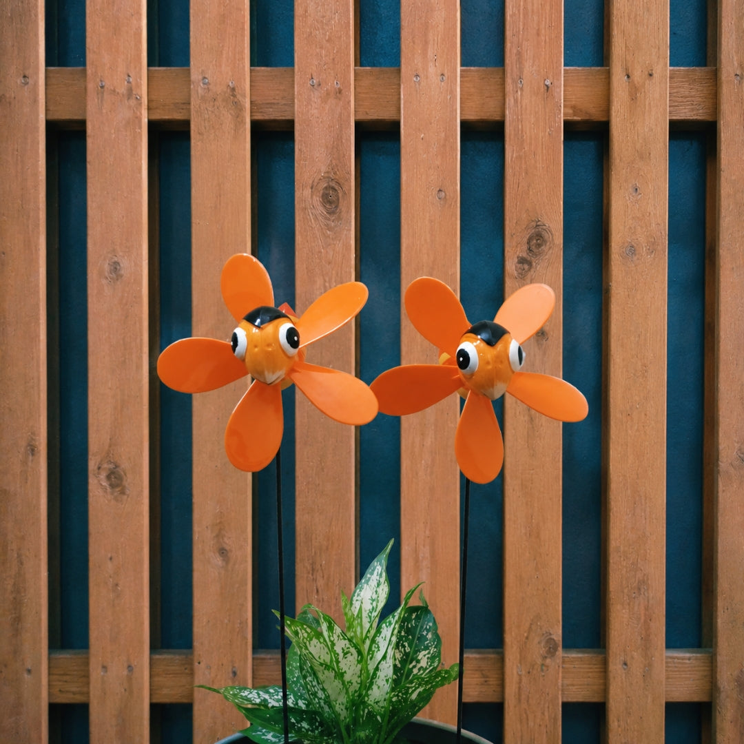 Two orange fish-shaped garden wind spinners with cartoon eyes mounted on black stakes, displayed in front of a brown wooden slat fence with a green plant at the bottom.
