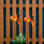 Two decorative garden wind spinner butterflies with orange and yellow striped bodies, black heads, and orange blades, mounted on stakes above a green plant in front of a wooden fence.