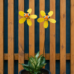 Two yellow flower-shaped garden wind spinners with smiling orange and black faces, mounted on stakes above a potted green plant in front of a wooden slat fence.