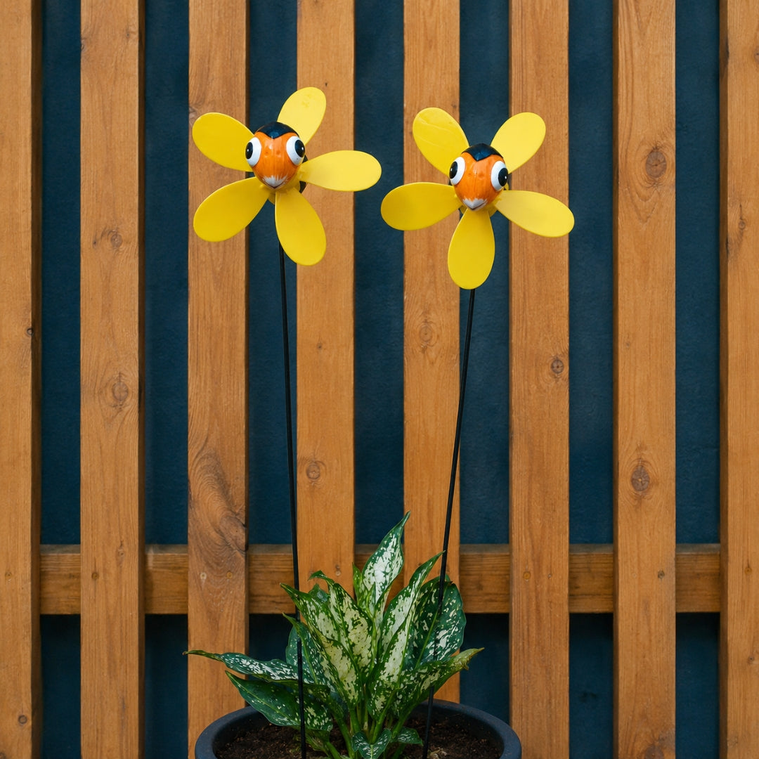 Two yellow flower-shaped garden wind spinners with smiling orange and black faces, mounted on stakes above a potted green plant in front of a wooden slat fence.
