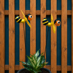 Two yellow and black bumblebee garden stakes with spinning wind-up fans, displayed in a black planter with a green variegated plant in front of a wooden fence and dark blue background.