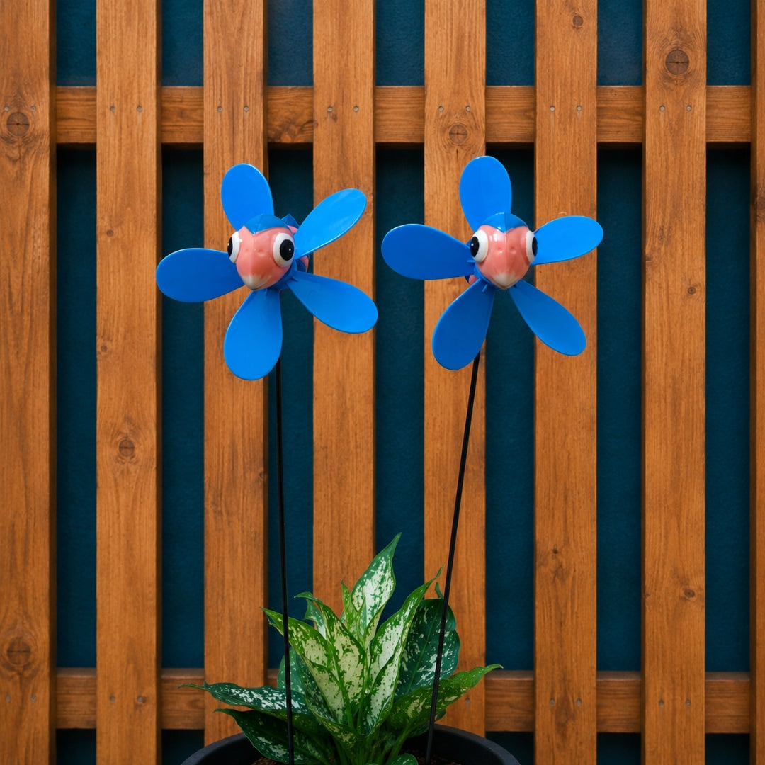 Two blue flower-shaped garden wind spinners with cartoon eyes mounted on stakes above a potted green plant, set against a wooden slat wall.