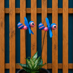 Two colorful metal garden stakes shaped like striped bumblebees with blue wings and pink-and-blue striped bodies, displayed in front of a wooden fence and above a potted green plant.