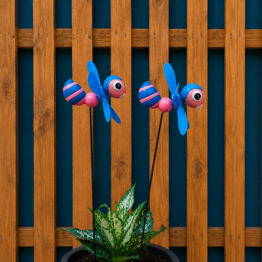 Two colorful metal garden stakes shaped like striped bumblebees with blue wings and pink-and-blue striped bodies, displayed in front of a wooden fence and above a potted green plant.