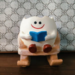 A small white ceramic figurine of a smiling tooth character sitting on a wooden rocking chair and holding a blue book, placed on a dark wooden surface against a patterned fabric background.