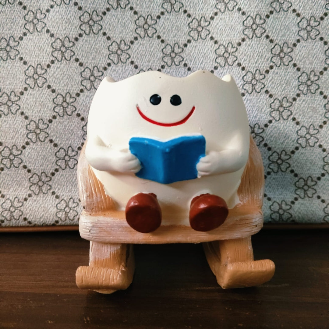 A small white ceramic figurine of a smiling tooth character sitting on a wooden rocking chair and holding a blue book, placed on a dark wooden surface against a patterned fabric background.