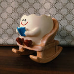 A ceramic tooth-shaped figurine sitting in a wooden rocking chair and reading a blue book, placed on a dark wooden surface against a patterned fabric background.
