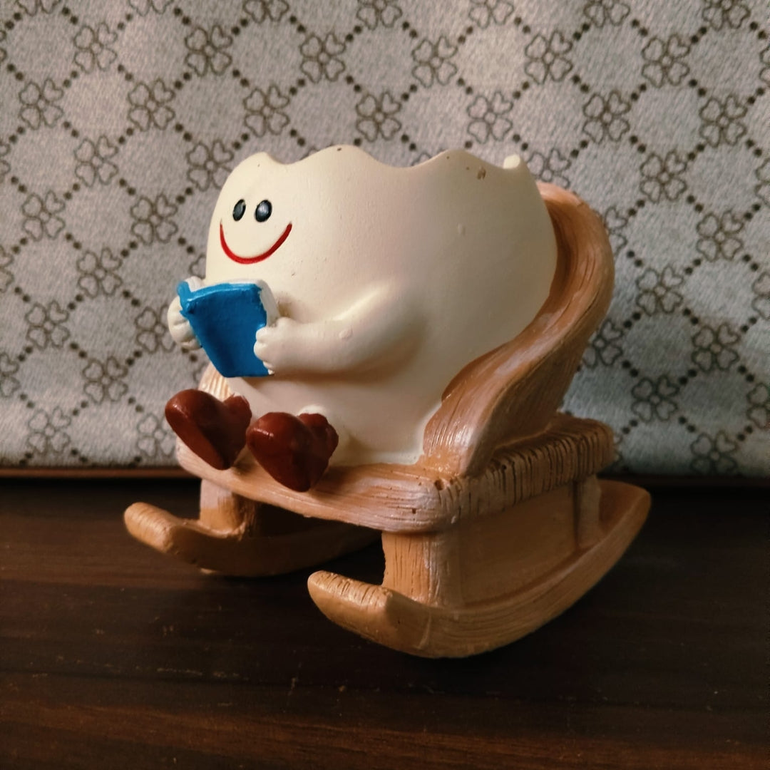 A ceramic tooth-shaped figurine sitting in a wooden rocking chair and reading a blue book, placed on a dark wooden surface against a patterned fabric background.