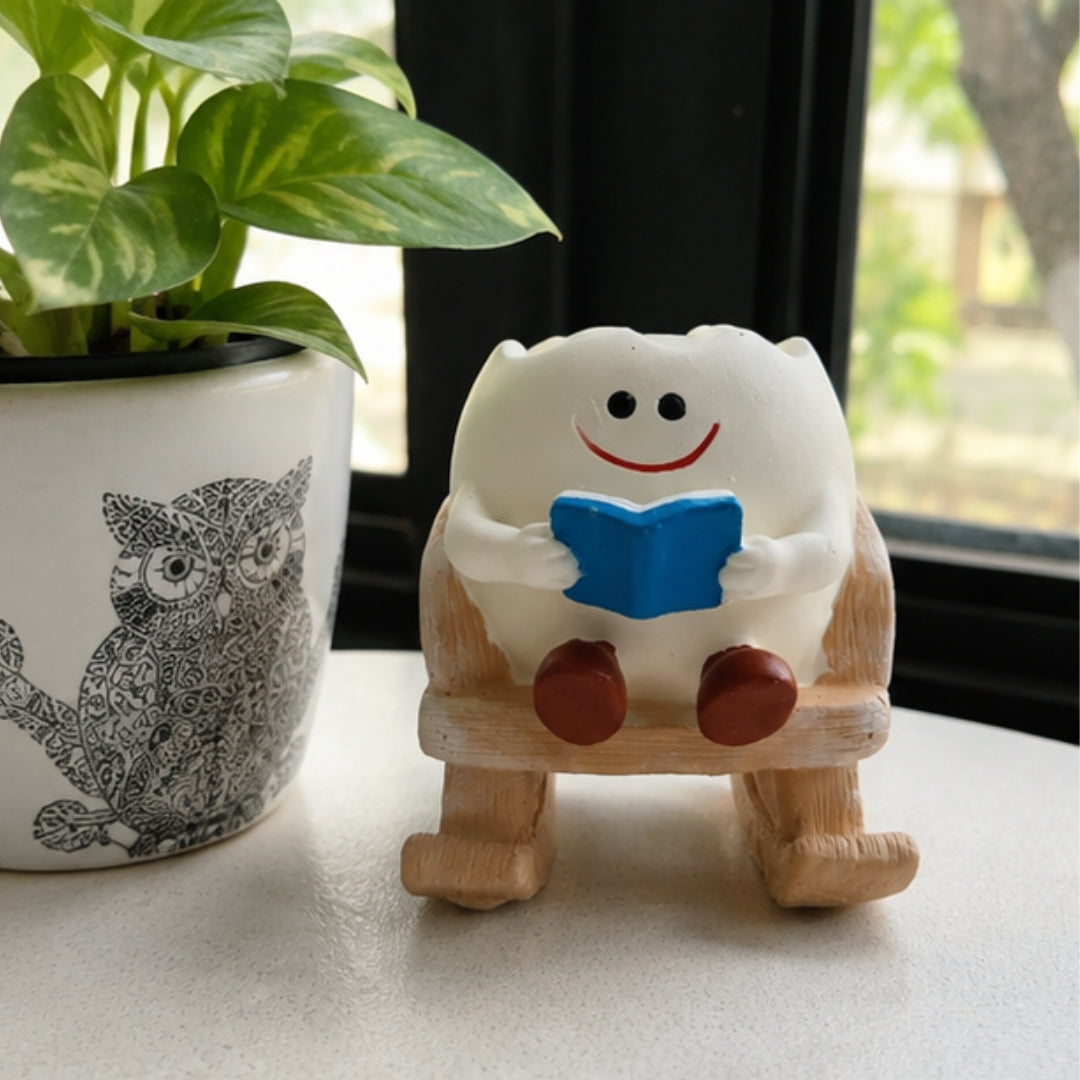 A small white cartoon figurine with a smiling face, black eyes, a red smile, and a blue book, sitting on a wooden rocking chair. The figurine is placed on a light surface next to a potted green plant.