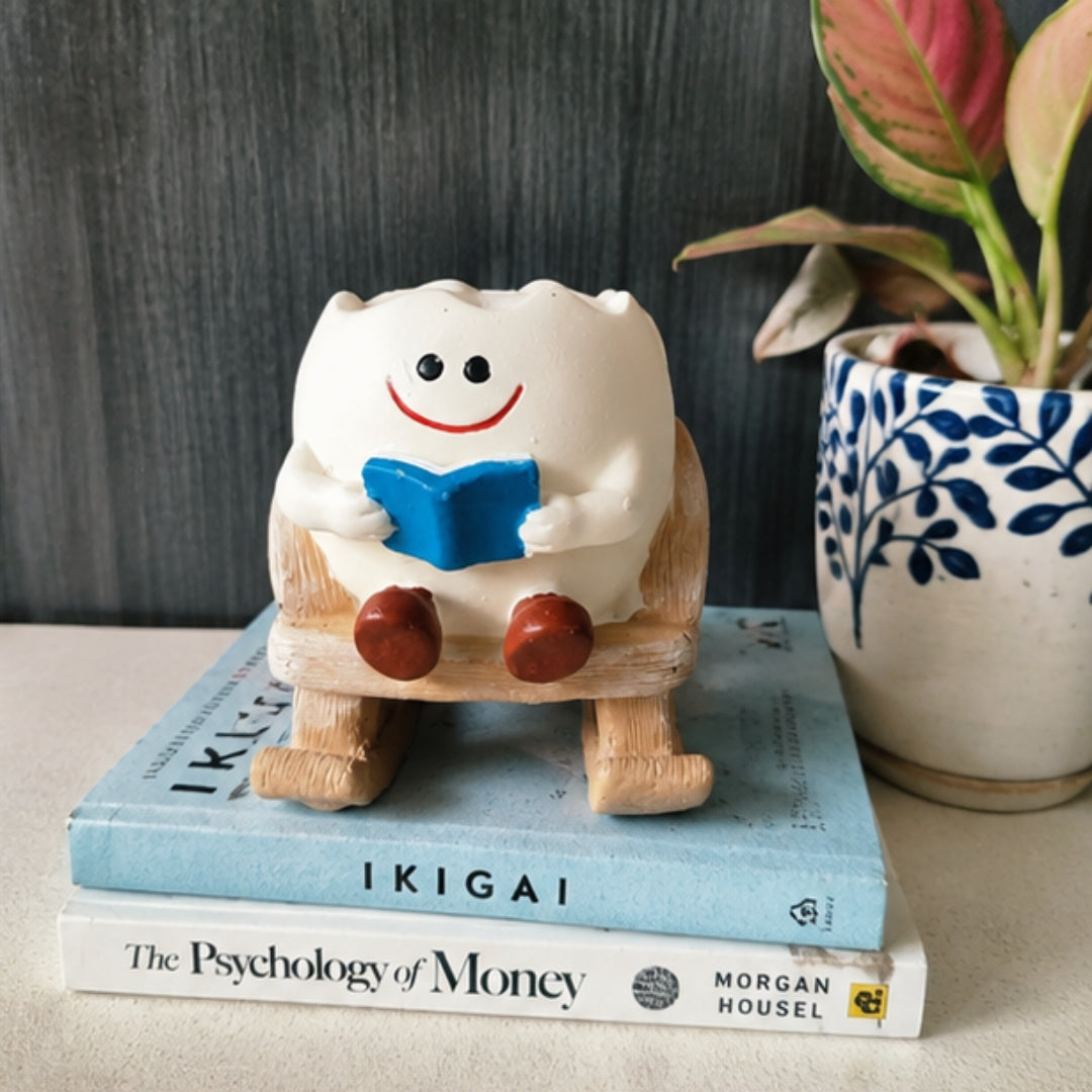 A small white ceramic tooth-shaped figurine with a smiling face, holding a blue book, sitting on a wooden rocking chair atop stacked books next to a potted plant.