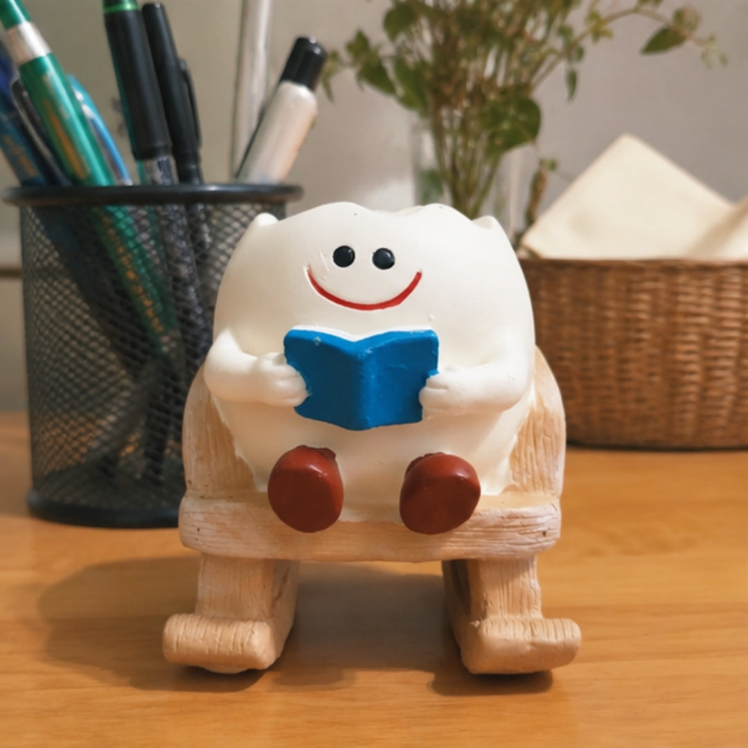 A small white cartoon character figurine sitting on a rocking chair and holding a blue book, placed on a wooden desk with a pen holder and basket in the background.