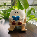 A small white cartoon figurine with a red smiley face sits on a wooden rocking chair, holding a blue book. The figurine is placed on a dark wooden table with green potted plants in the background.