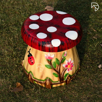 Mushroom-shaped stool with red cap and white spots, beige body with floral and ladybug design, on grass.