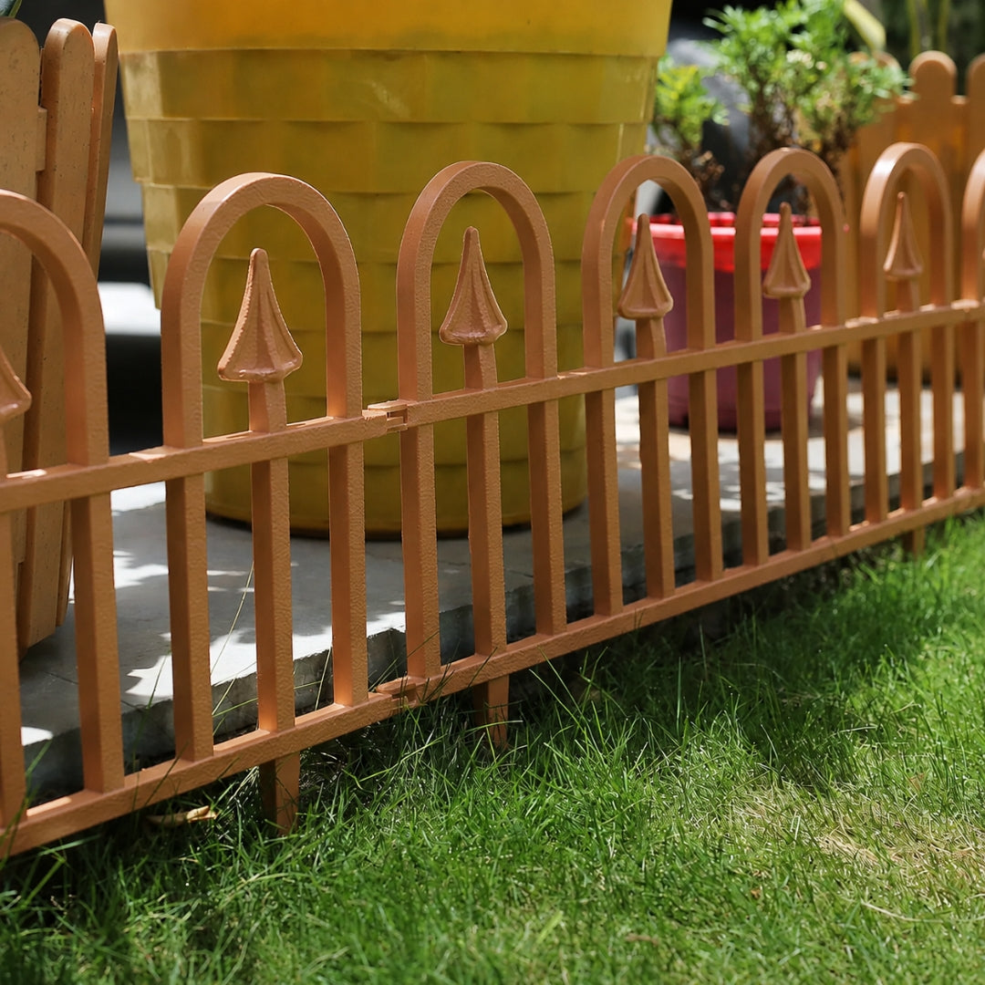 A close-up of a decorative metal picket fence in a warm coppery-brown finish, installed outdoors on a stone surface with green grass in the foreground and a large yellow planter in the background.