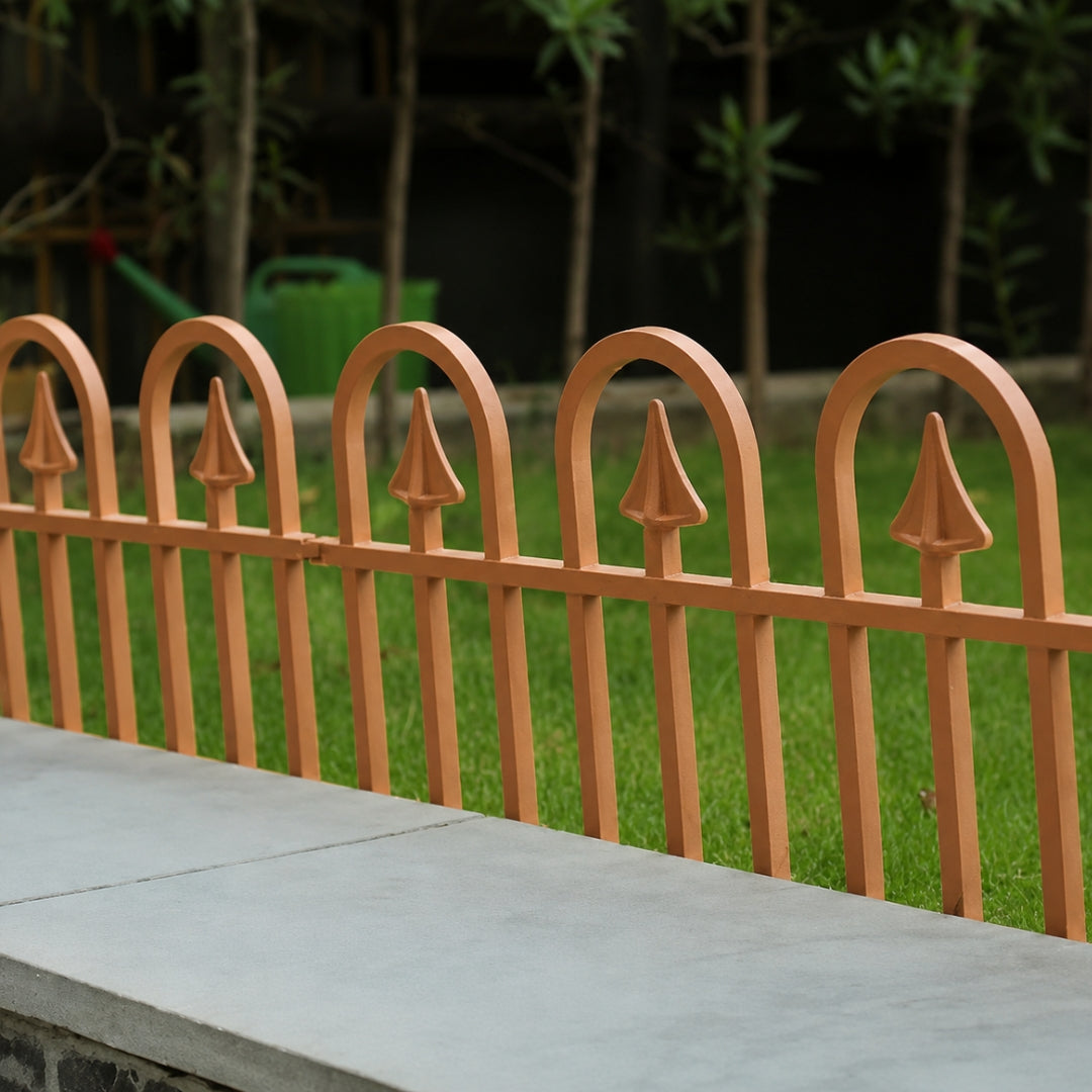 A close-up of a light brown metal garden fence with arched tops and pointed finials, installed along a concrete ledge with green grass and trees in the background.