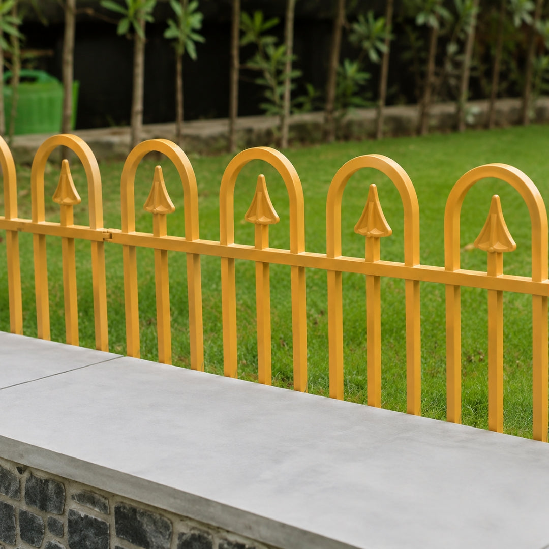 Yellow metal garden fence with arched picket tops mounted on a stone wall behind a concrete ledge, with green grass and trees in the background.