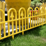 A bright yellow metal decorative fence with pointed tops and arched vertical bars, shown outdoors on green grass in front of a large yellow planter.
