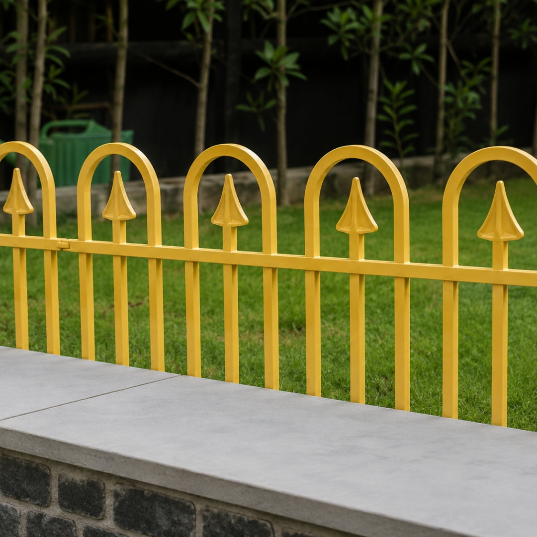 Yellow metal decorative fence with arched tops and pointed finials, shown outdoors behind a gray concrete ledge with green grass and trees in the background.