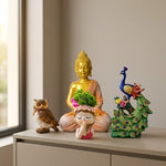 A decorative tabletop arrangement featuring a gold Buddha statue, a colorful peacock figurine, a small owl figurine, and a praying girl planter with green foliage, displayed on a light beige cabinet in a softly lit interior.