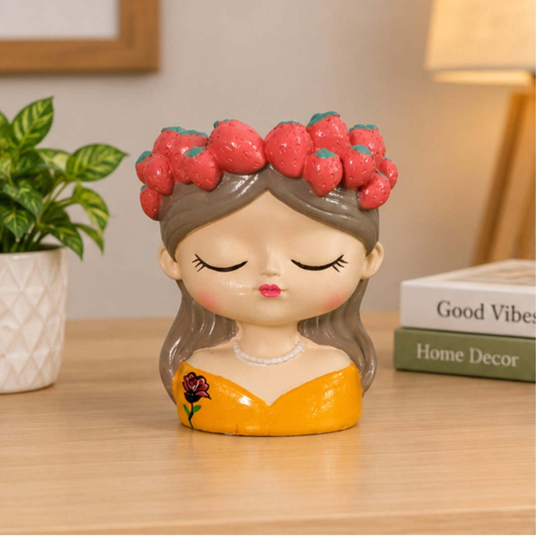 A decorative ceramic figurine of a sleeping girl with brown hair, a strawberry crown, and a yellow dress, displayed on a wooden table next to a potted plant and books.