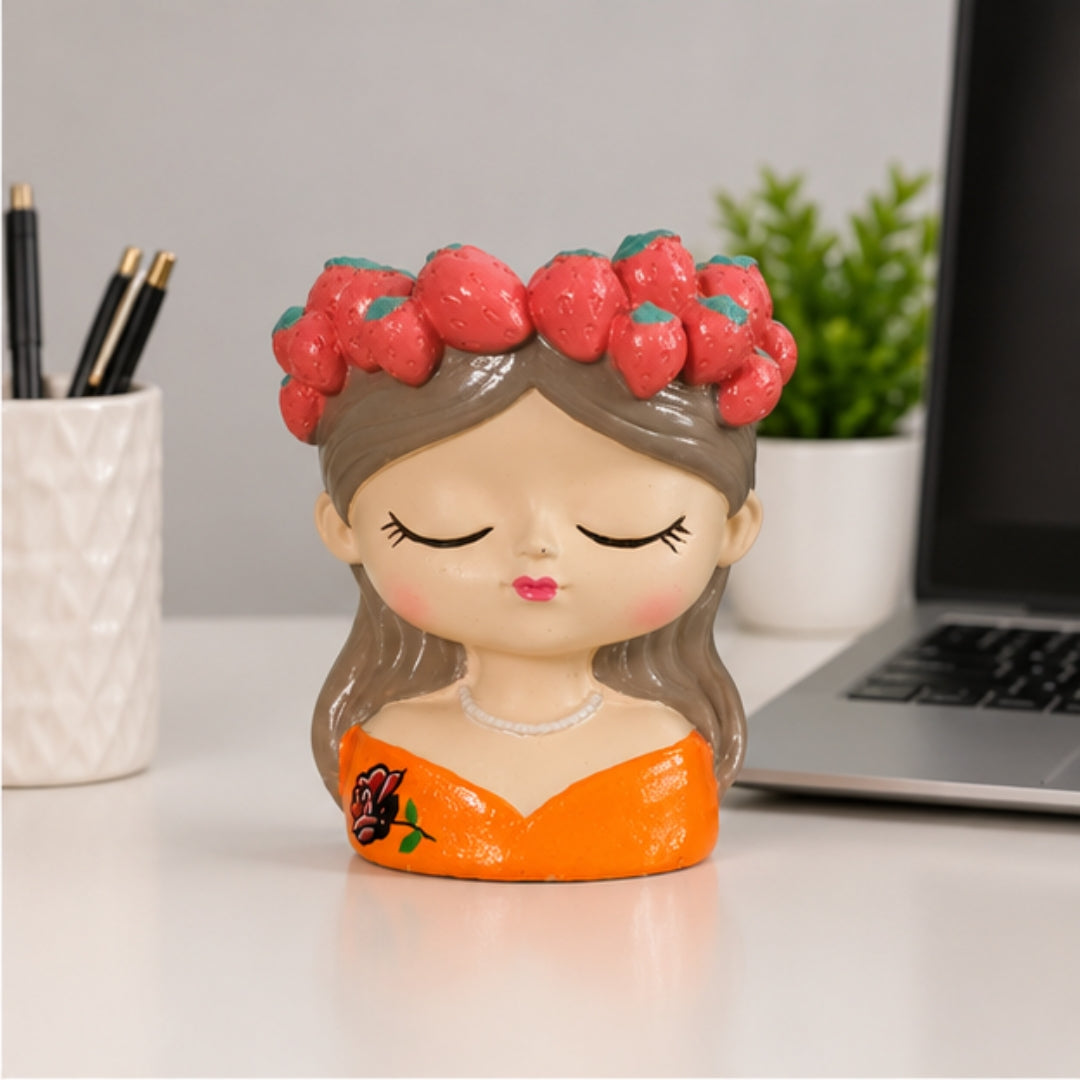 A ceramic figurine of a sleeping girl with brown hair, a red strawberry crown, and an orange dress, placed on a white desk next to a laptop and a plant.