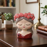 A decorative ceramic figurine of a sleeping girl with closed eyes, wearing a pink strawberry flower crown and a red dress, displayed on a wooden table in a home setting.