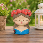 A decorative ceramic figurine of a sleeping girl with brown hair, a red strawberry crown, and a blue dress, displayed on a wooden table with a potted plant and a lit lantern in a garden setting.