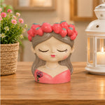 A decorative figurine of a sleeping girl with closed eyes, wearing a pink strawberry crown and a pink dress, placed on a wooden table with a potted plant and a white lantern in the background.