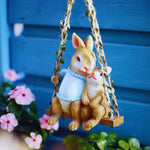 Cute bunny figurines on a swing, garden decor with flowers and blue background