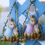 Cute bunny figurines in sweaters sitting on swings, outdoor garden decor against blue lattice.