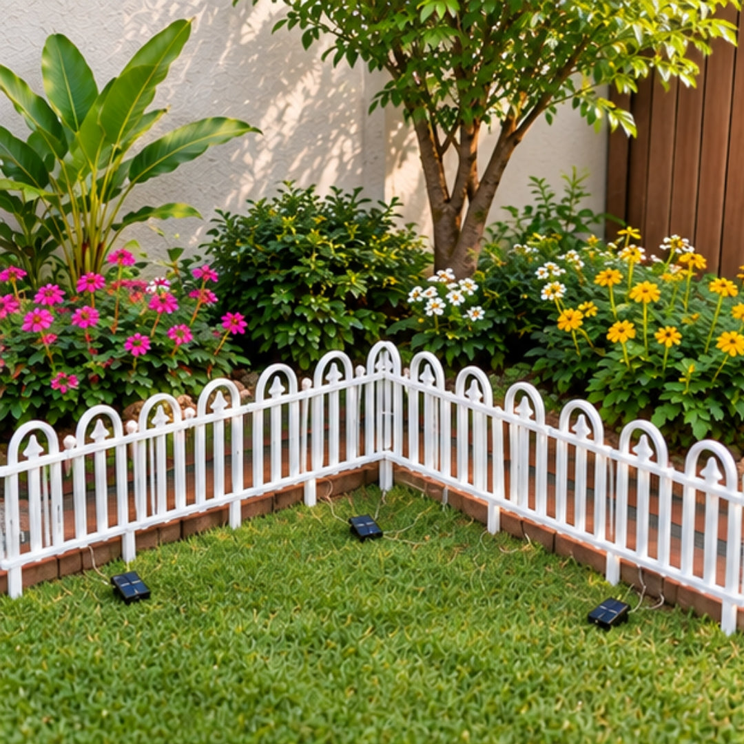 A white decorative picket fence with solar-powered lights in a green garden corner with flowers and plants.
