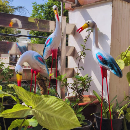Decorative storks in a garden setting with plants and a building in the background.