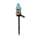 A miniature fairy garden stake with a small cottage featuring a blue flower-covered roof, a brown door, and a black solar-powered stake with an attached solar panel, shown on a plain white background.