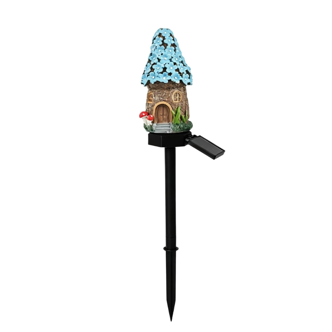 A miniature fairy garden stake with a small cottage featuring a blue flower-covered roof, a brown door, and a black solar-powered stake with an attached solar panel, shown on a plain white background.