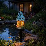 A decorative fairy-tale house garden stake light with a glowing warm yellow body, blue flower-like roof, and small solar panel, photographed outdoors beside a pond at night.