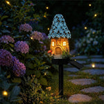 A decorative garden fairy light shaped like a small cottage with a blue flower-covered roof and warm glowing windows, mounted on a black solar-powered stake in a garden at night.