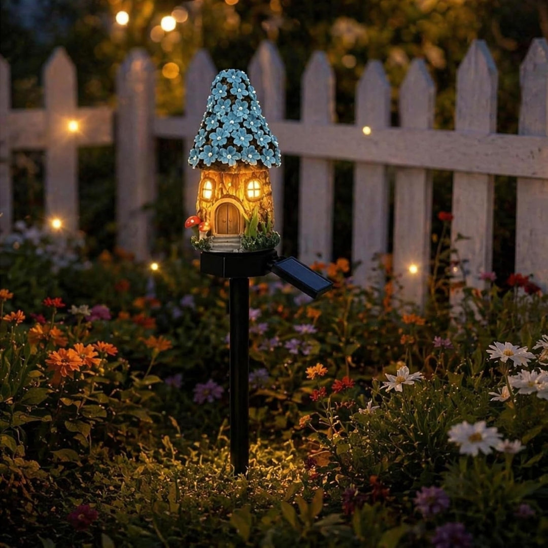 A decorative miniature house garden stake with a glowing lit house design, blue flower-patterned roof, and a small solar panel attached to the stake, set in a garden bed at night in front of a white picket fence.