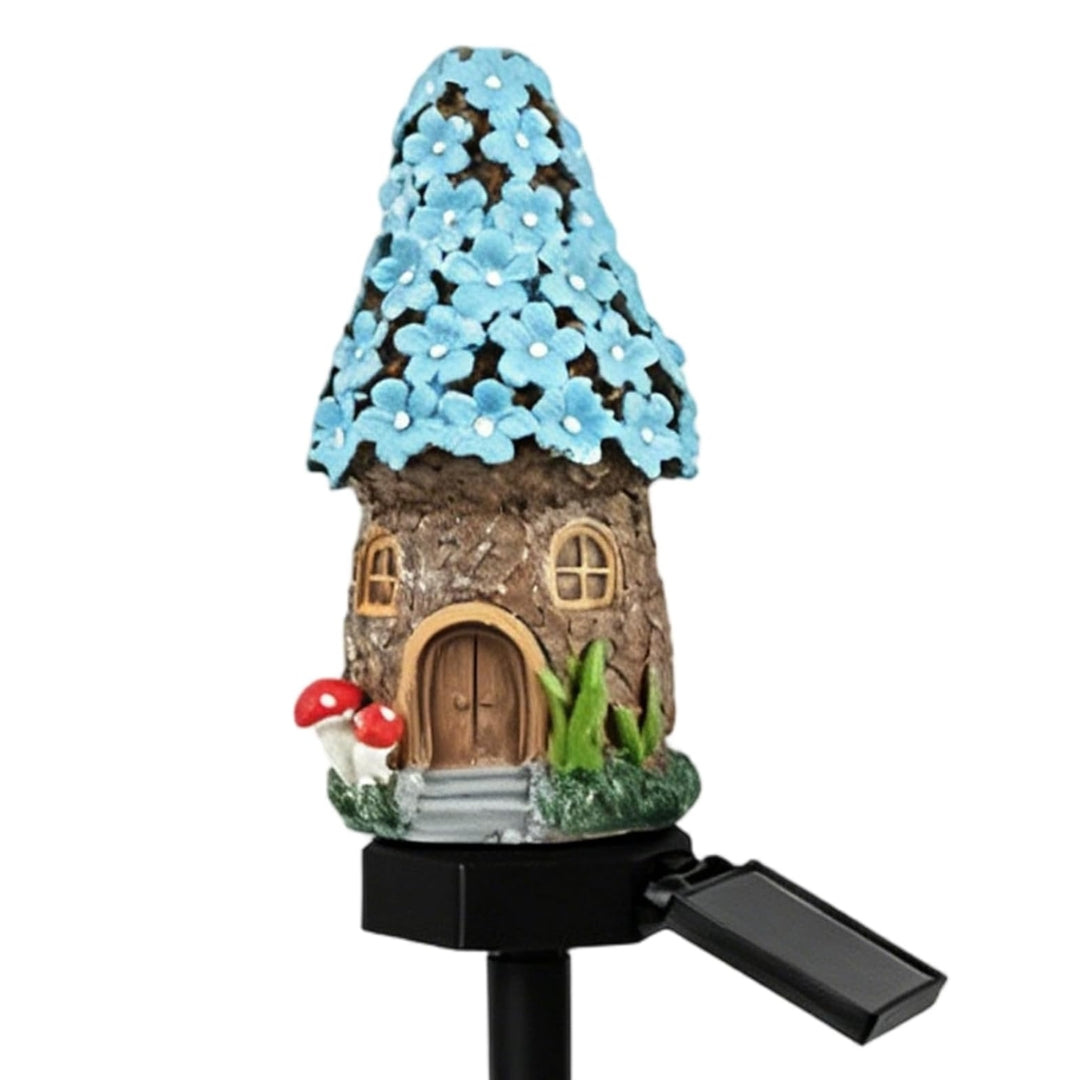 A miniature fairy house garden stake with a brown textured body, a blue flower-covered roof, red-and-white mushrooms, green grass, and a black solar-powered stake with a small solar panel extending to the right, shown on a plain white background.