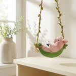 A pink pig figurine reclining in a green swing seat suspended by twine with leafy vines, displayed on a white table in a bright indoor setting.